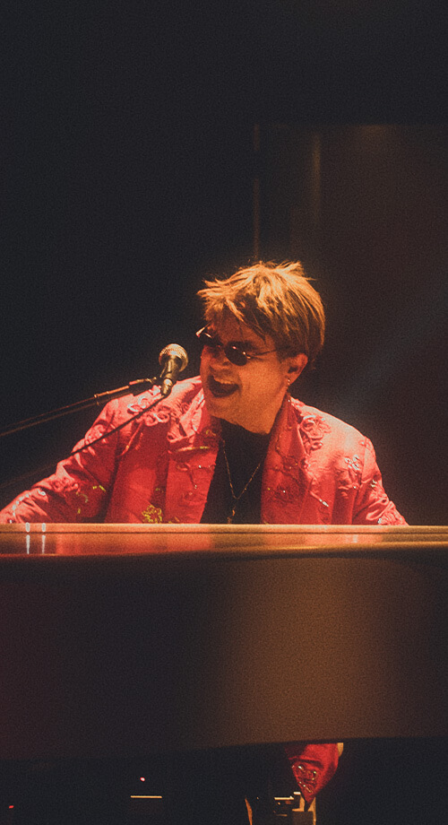 Rogério Martins caracterizado de Elton John tocando piano no show Your Song.